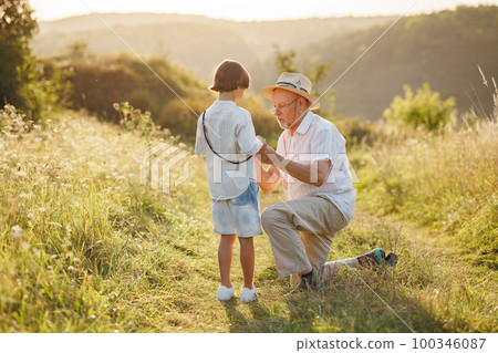 Backside photo of little boy and his grandfather standing and talking in the field. Boy and grandfather spending time together at summer. Brunette boy and man wearing striped shirts. 100346087