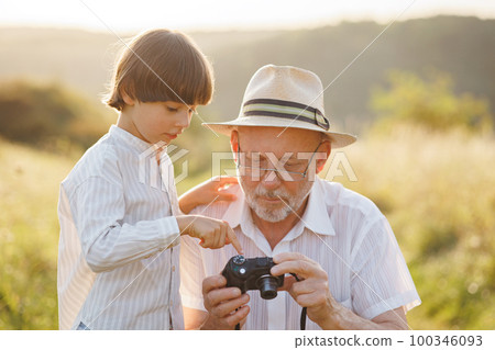 Portrait of little explorer and his grandfather with camera in a field. Boy and grandfather spending time together at summer. Brunette boy and man wearing striped shirts. 100346093