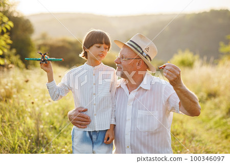 Portrait of little explorer and his grandfather playing with two toys plane in a field. Boy and grandfather spending time together at summer. Brunette boy and man wearing striped shirts. 100346097
