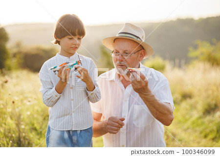 Portrait of little explorer and his grandfather playing with two toys plane in a field. Boy and grandfather spending time together at summer. Brunette boy and man wearing striped shirts. 100346099