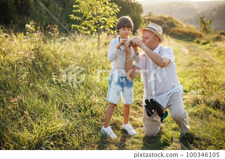 Portrait of little explorer and his grandfather with binoculars in a field. Boy and grandfather spending time together at summer. Brunette boy and man wearing striped shirts. 100346105