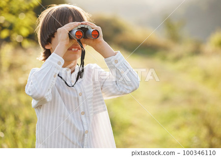 Portrait of little explorer with binoculars in a field. Little caucasian boy looking through a binoculars in a field at summer. Brunette boy wearing striped shirt. 100346110