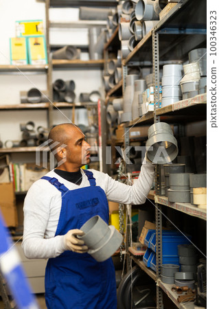 Salesman in overalls with plumbing fittings at hardware store warehouse 100346323