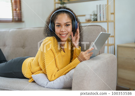 Portrait of an African American sitting on a sofa using tablet and wearing headphones to relax. 100353586