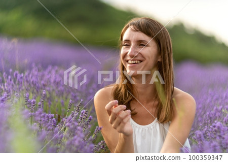 Close up portrait of happy young brunette woman in white dress on blooming fragrant lavender fields with endless rows. Warm sunset light. Bushes of lavender purple aromatic flowers on lavender fields. Close up portrait of happy young brunette woman in white dress on blooming fragrant lavender fields with endless rows. Warm sunset light. Bushes of lavender purple aromatic flowers on lavender fields. 100359347