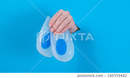 Woman holding orthopedic silicone heel pads. A woman's hand sticks out of a hole in a cardboard blue background. 100359482