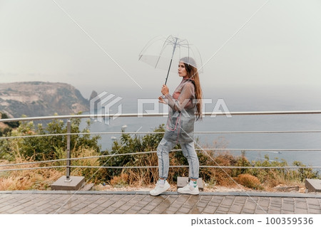 Woman rain umbrella. Happy woman portrait wearing a raincoat with transparent umbrella outdoors on rainy day in park near sea. Girl on the nature on rainy overcast day. Woman rain umbrella. Happy woman portrait wearing a raincoat with transparent umbrella outdoors on rainy day in park near sea. Girl on the nature on rainy overcast day. 100359536