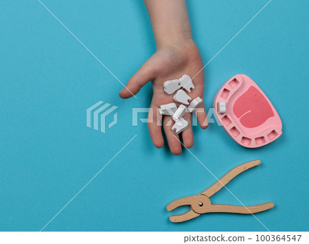 Dentist game. A child's hand holds toy wooden teeth. A toy jaw without teeth and forceps for removal nearby. 100364547