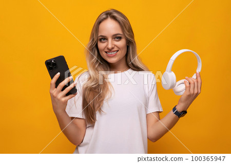 a girl with a smartphone and headphones in her hands uses an application on an orange background 100365947