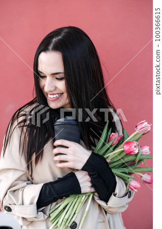 Spring outdoor portrait of happy woman with tulips bouquet and coffee to go on spring city street. Happy young woman holding bouquet of pink flowers 100366615