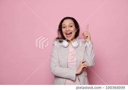 Amazed multi-ethnic woman in wireless headphones, pointing index finger up at copy advertising space for text, expressing idea and finding solution, posing with open mouth on isolated pink background Amazed multi-ethnic woman in wireless headphones, pointing index finger up at copy advertising space for text, expressing idea and finding solution, posing with open mouth on isolated pink background 100368099