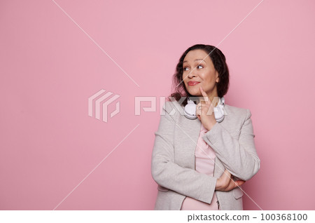 Pensive middle-aged pretty woman with wireless headphones, dreamily looking aside at copy space for advertising text, posing with arms crossed, holding her finger on chin over isolated pink background 100368100