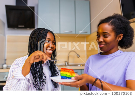 Beautiful woman feeding her wife with rainbow colorful cake and celebrating anniversary or Valentine 100370108