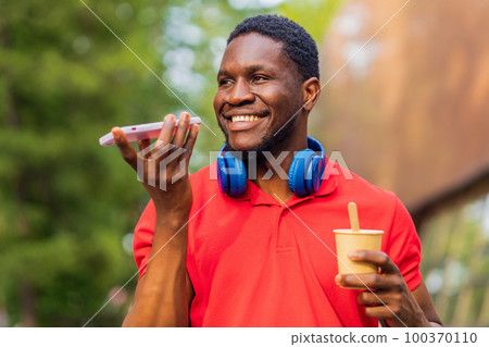 young afro american man with headphones on neck using smartphone in summer park 100370110