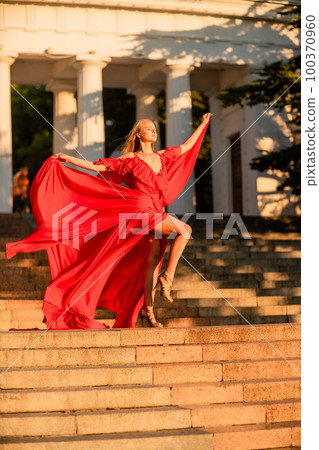 A woman in a long red dress against the backdrop of sunrise, bright golden light of the sun's rays. The concept of femininity, harmony. 100370960