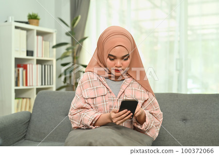 Charming young Muslim woman in hijab typing massage on her mobile phone while sitting on couch at home Charming young Muslim woman in hijab typing massage on her mobile phone while sitting on couch at home 100372066