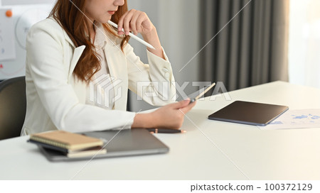 Concentred milennila female corporate CEO in elegant suit using digital tablet at her workplace 100372129