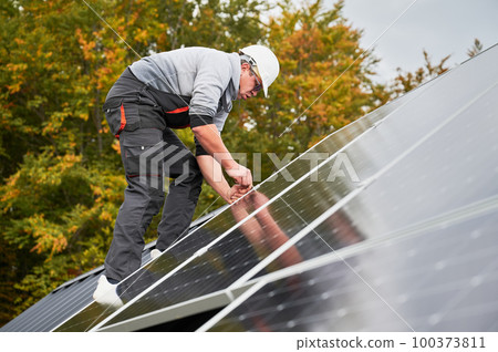 Man electrician mounting photovoltaic solar panels on roof of house. Engineer in helmet installing solar module system with help of hex key. Concept of alternative, renewable energy. 100373811