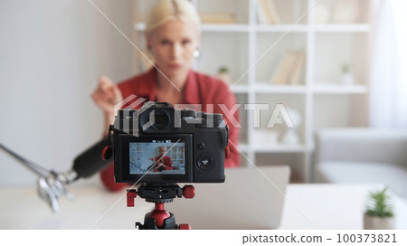 Influencer vlog. Live streaming. Online coaching. Mature blogger woman talking recording video interview on camera on tripod on defocused interior with free space. Influencer vlog. Live streaming. Online coaching. Mature blogger woman talking recording video interview on camera on tripod on defocused interior with free space. 100373821