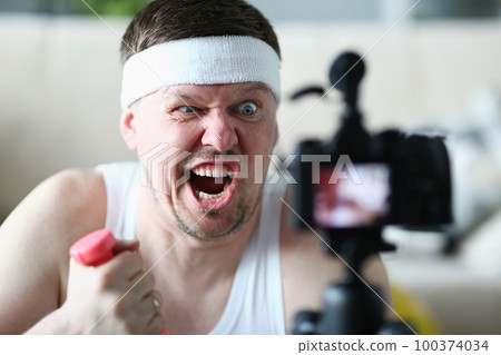 Portrait of an emotionally male blogger with grimace on face and holding dumbbells in front of camera 100374034
