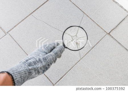 The hand of an industrial worker in a construction glove with a magnifying glass examines the damaged cracked tile background. Renovation concept The hand of an industrial worker in a construction glove with a magnifying glass examines the damaged cracked tile background. Renovation concept 100375392