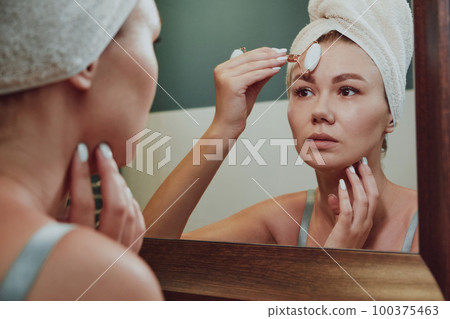 Close up woman using jade facial roller for face massage looking in the bathroom mirror 100375463