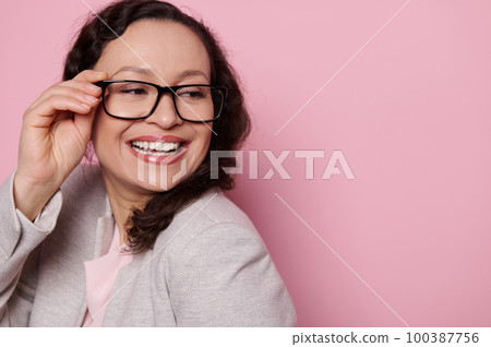 Close-up portrait of a happy middle-aged pretty woman, a positive teacher in classic light gray suit and stylish glasses, smiling broadly, looking aside over isolated pink background with copy space. Close-up portrait of a happy middle-aged pretty woman, a positive teacher in classic light gray suit and stylish glasses, smiling broadly, looking aside over isolated pink background with copy space. 100387756