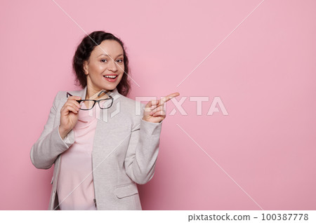 Portrait of African American middle-aged successful confident woman in light gray elegant suit, holding stylish trendy glasses and looking at camera, pointing at copy space on isolated pink background Portrait of African American middle-aged successful confident woman in light gray elegant suit, holding stylish trendy glasses and looking at camera, pointing at copy space on isolated pink background 100387778
