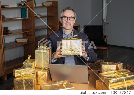 Mature happy smilimg  businessman in glasses and blue suit with laptop and a lot of gifts around.  100387847