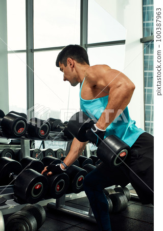 Man doing exercise with dumbbell leaning on sports bench in the gym. Photo of a sexy muscular man in sportswear and good physique on grey background. Strength and motivation, sport, fitness goal 100387963