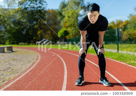 Asian sportsman tired after running and active physical exercise, man taking breath and rest on sunny day in stadium, overtired after running. Asian sportsman tired after running and active physical exercise, man taking breath and rest on sunny day in stadium, overtired after running. 100388909