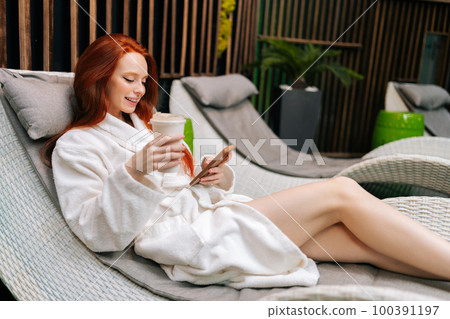 Cheerful young woman in white bathrobe relaxing with mobile sitting on deck chair by poolside and drinking cocktail after beauty treatment at spa salon. Concept of wellness spa resort, cosmetology. 100391197