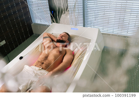 High-angle view of handsome bearded young man lying relaxing on electric massage bed and enjoying spa treatment at luxury spa salon. Muscular male having rest and enjoying wellness weekend alone. 100391199
