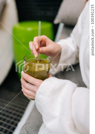 Vertical closeup cropped shot of unrecognizable young woman in white bathrobe holding glass with delicious healthy freshly squeezed vitamin juice in hand, sitting on lounger at spa salon. Vertical closeup cropped shot of unrecognizable young woman in white bathrobe holding glass with delicious healthy freshly squeezed vitamin juice in hand, sitting on lounger at spa salon. 100391235