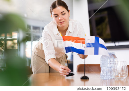 Positive young woman putting flag of Holland on the table with flag of Finland in conference room 100392452