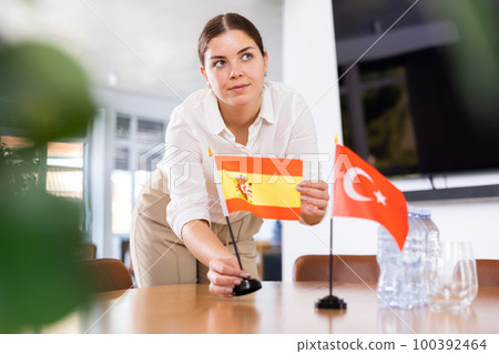 Young female assistant putting up turkish and spanish flags on table for international negotiations 100392464