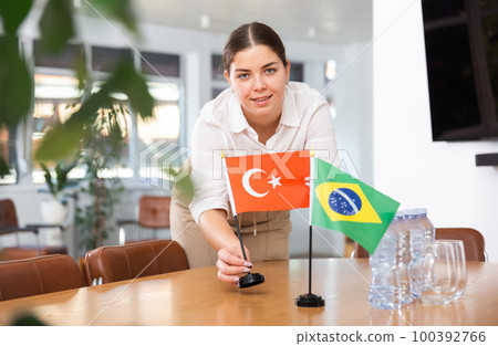 Young female assistant putting up brazilian and turkish flags on table for international negotiations 100392766