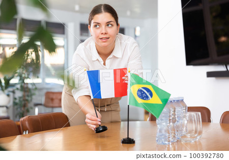 Positive young woman putting flag of France on the table with flag of Brazil in conference room 100392780