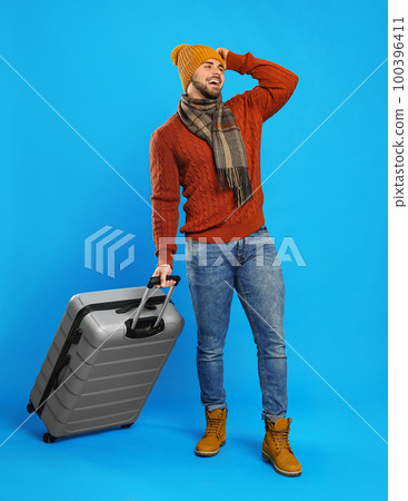 Young man in warm clothes with suitcase on blue background. Winter vacation 100396411