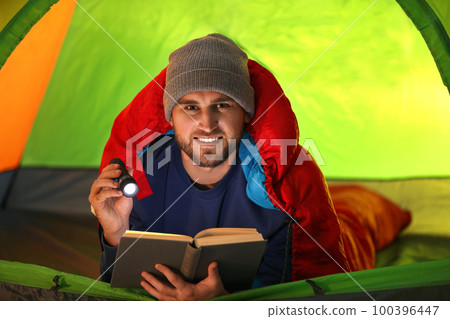 Young man with flashlight reading book in tent 100396447