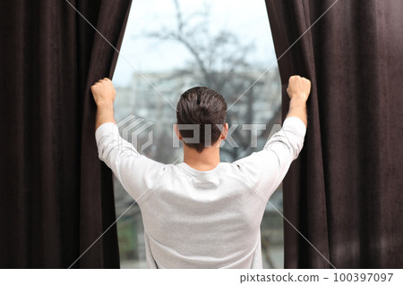Man opening window curtains at home, back view Man opening window curtains at home, back view 100397097