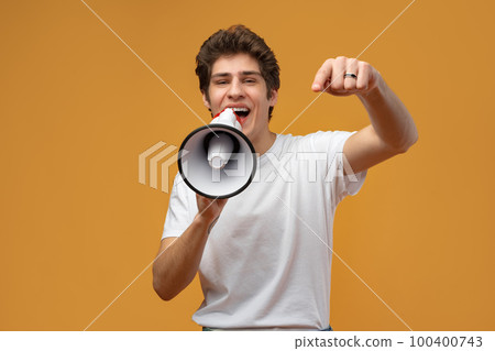 Young man shouting into megaphone making announcement against yellow background Young man shouting into megaphone making announcement against yellow background 100400743