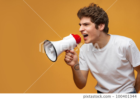 Young man shouting into megaphone making announcement against yellow background 100401344