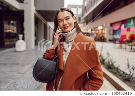 Cute brunette woman in brown coat talking on the phone in the city Cute brunette woman in brown coat talking on the phone in the city 100402419