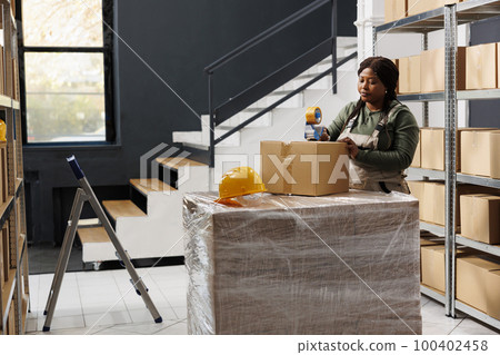 Stockroom supervisor preparing customers orders, putting adhesive tape on cardboard box before shipping packages. African american employee working at merchandise inventory in warehouse 100402458