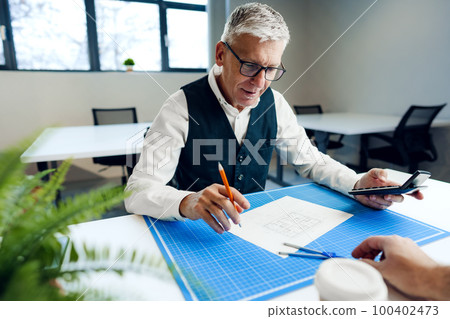 Busy senior man sitting at office desk with papers and making notes Busy senior man sitting at office desk with papers and making notes 100402473