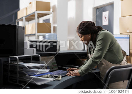 Storehouse manager analyzing goods logistics on laptop computer, working at storage room inventory typing report. African american employee preparing customers orders in warehouse Storehouse manager analyzing goods logistics on laptop computer, working at storage room inventory typing report. African american employee preparing customers orders in warehouse 100402474