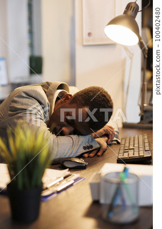 Tired executive manager with burnout syndrome sleeping at work on desk because of extreme fatigue. Exhausted businessman suffering from sleepiness after working late at night at company project 100402480