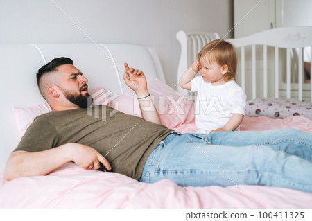 Strict father young man and baby girl little daughter on bed in room at home Strict father young man and baby girl little daughter on bed in room at home 100411325