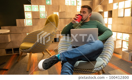 Caucasian bearded man sits in a comfortable chair drinks coffee and works on a laptop. Freelancer.  100412800